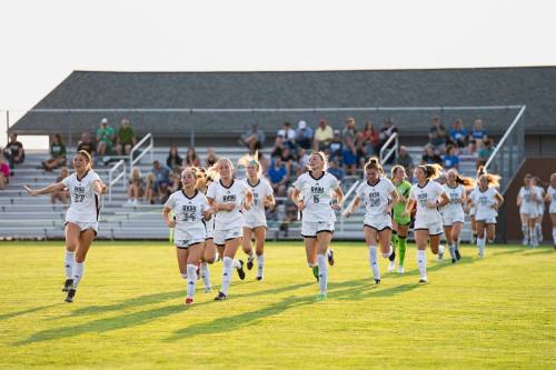 The soccer team runs out onto a green field, spectators watch from the stands in the background
